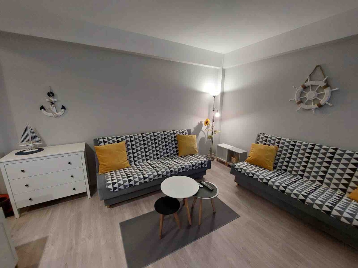 A modern living area is designed with two patterned sofas and a small round coffee table. Soft yellow cushions add a pop of color. A light source from a floor lamp illuminates the space, while an anchor and nautical decor piece are displayed on the wall.