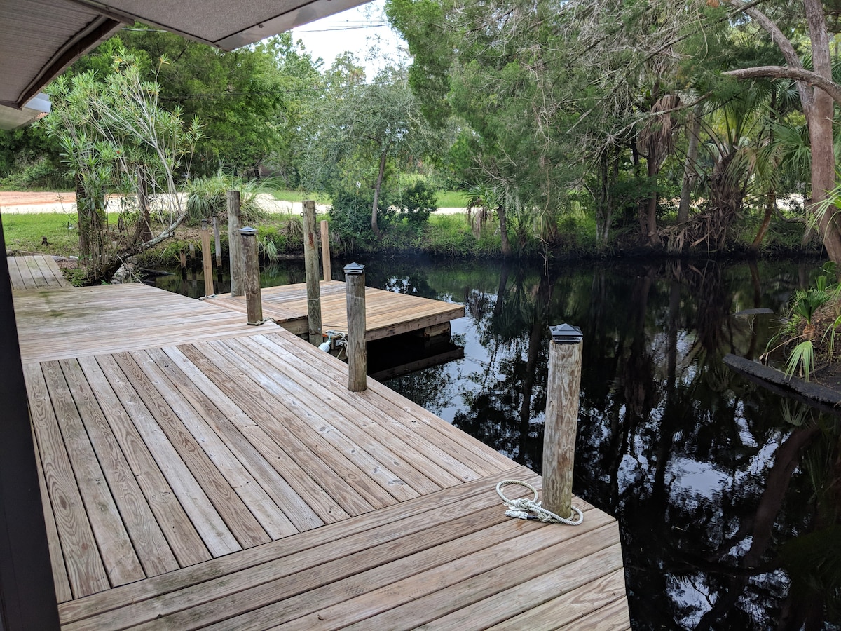 A wooden dock extends over calm waters, surrounded by lush greenery. Posts line the edge of the dock, providing a structured environment for recreational activities. Dense foliage frames the scene, enhancing the natural setting.