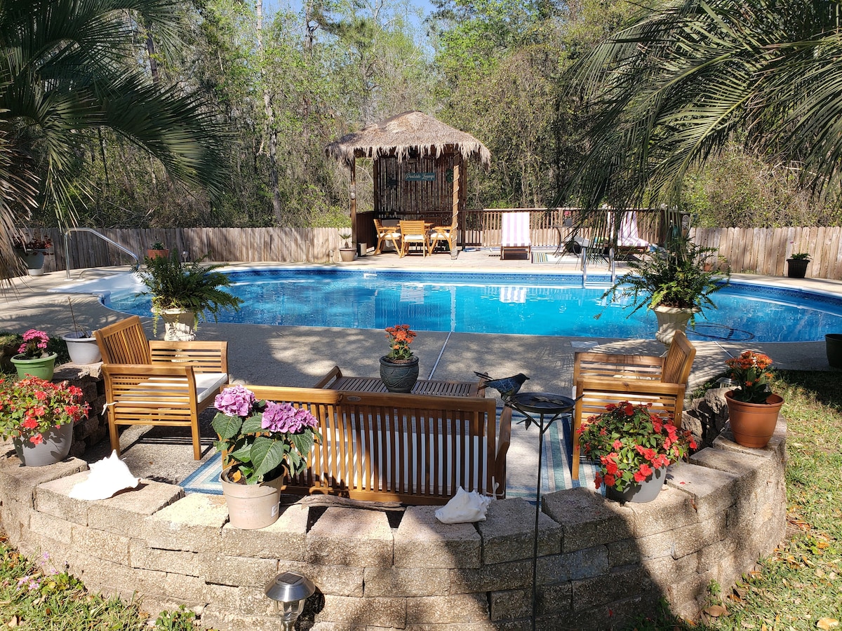 A serene pool area is framed by lush greenery and decorative plants. Comfortable seating options are positioned around a circular stone border, while a tiki-style hut offers a shaded retreat. The bright blue water is inviting, harmonizing with the natural surroundings.