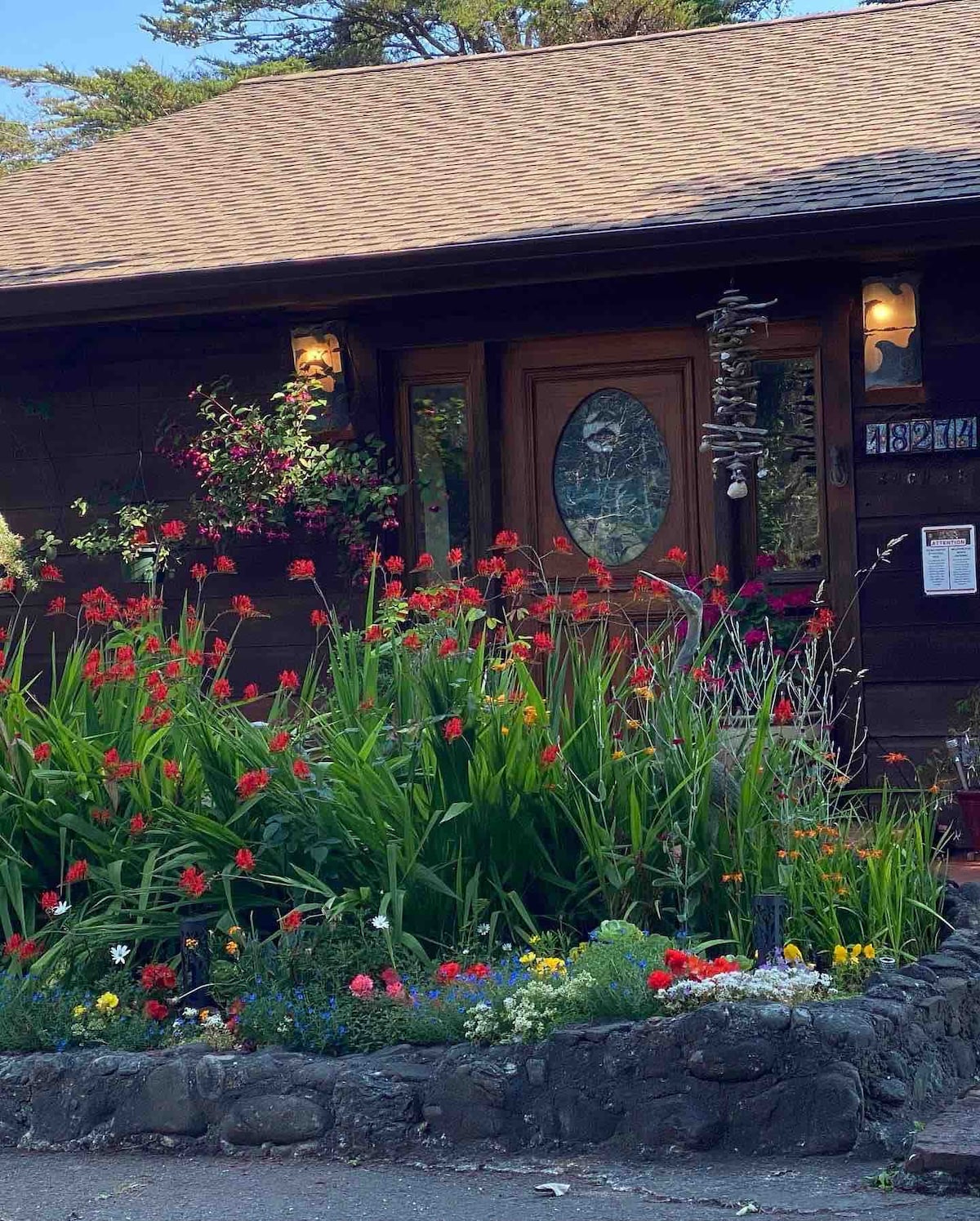 The entrance of a rustic wooden cottage is framed by vibrant flower beds, with various colorful blooms and lush green foliage. The front door features a decorative glass design and is flanked by welcoming wall sconces, creating an inviting ambiance.