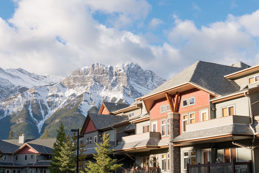 A scenic view showcases the exterior of the Cougar Lodge at The Lodges at Canmore, featuring a mix of wood and stone architecture. Majestic snow-capped mountains serve as a backdrop, with clear blue skies and a few clouds enhancing the natural beauty of the location.