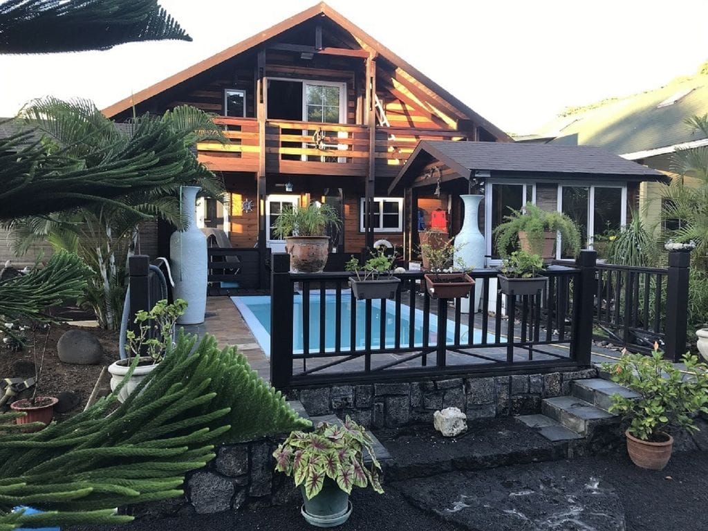 The chalet features a wooden exterior with a steeply pitched roof and large balcony. A private pool is situated at the front, surrounded by a black fence and potted plants. Lush greenery and palm trees offer a sense of seclusion, complementing the inviting outdoor space.