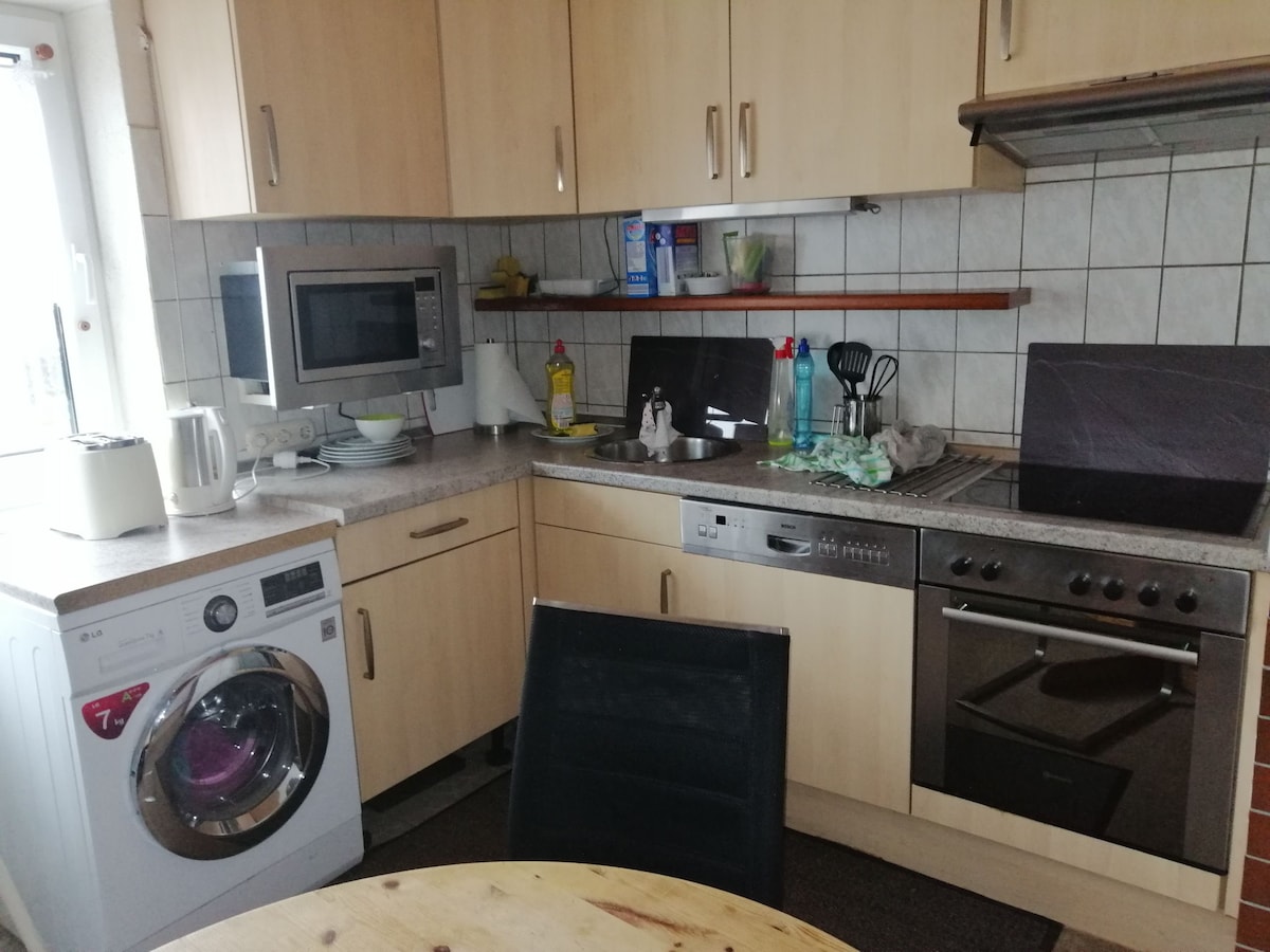 A functional kitchen is featured with light wooden cabinetry and a tiled backsplash. Essential appliances include a microwave, a stove, and a washing machine. A round dining table is situated in the foreground, accompanied by a black chair, creating a practical cooking and dining area.