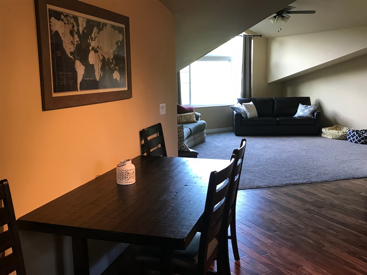 A dining area features a simple wooden table surrounded by four chairs. A world map hangs on the wall nearby. The spacious living area is visible beyond, showcasing a large window and a sofa with decorative cushions.