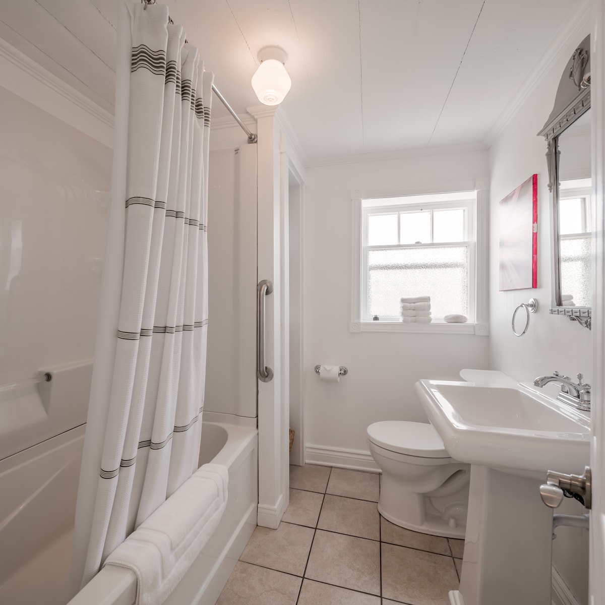 The bathroom features a clean design with a bathtub/shower combination, complemented by a white shower curtain with subtle stripes. A pedestal sink and a toilet are present, with bright lighting from a ceiling fixture and natural light filtering through a window adorned with neatly folded towels.
