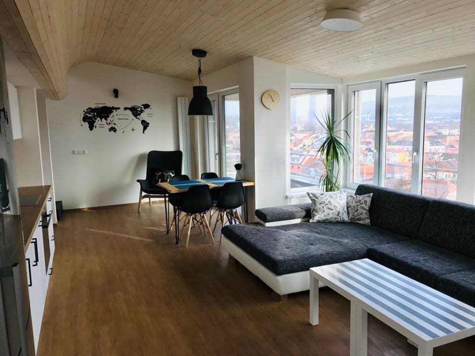 The spacious living area features large windows offering panoramic city views. A comfortable sectional sofa in gray is positioned next to a modern dining table with dark chairs. A world map accent decorates the wall, and a plant adds a touch of greenery.