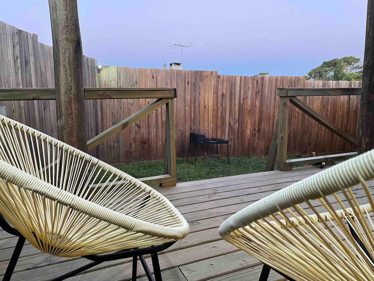 Two comfortable chairs are positioned on a wooden deck, inviting relaxation. A fenced yard extends beyond, with lush grass visible. The wooden fence adds privacy to the outdoor space, creating a serene setting for guests to enjoy.