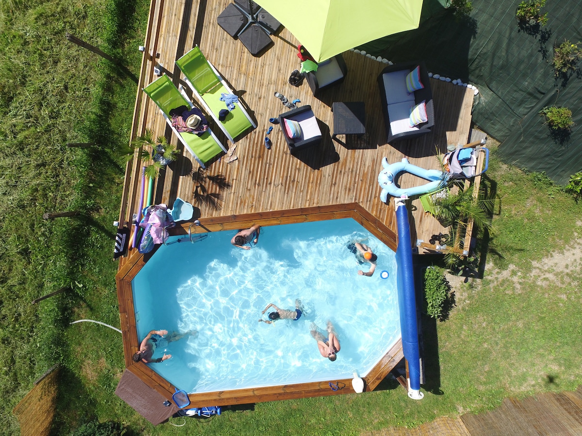 An overhead view of a landscaped wooden deck surrounding a rectangular pool. People are seen enjoying the water. Lounge chairs with green cushions and a shaded seating area are positioned nearby, with vibrant plants enhancing the outdoor setting.