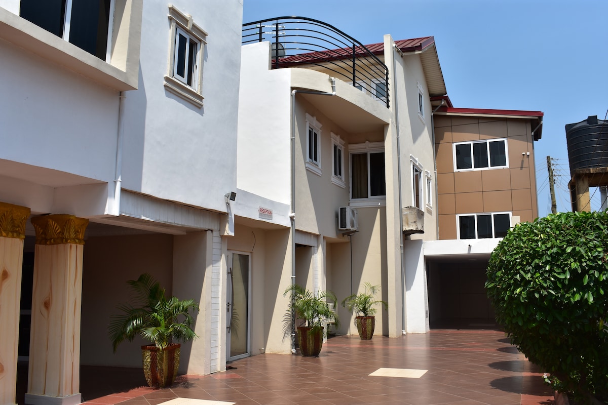 A well-maintained exterior view of the property is displayed, showcasing modern architectural elements. Potted plants are arranged along the pathway, with a mix of textured walls and large windows reflecting the sunlight. A spacious, paved area promotes ease of access and movement.