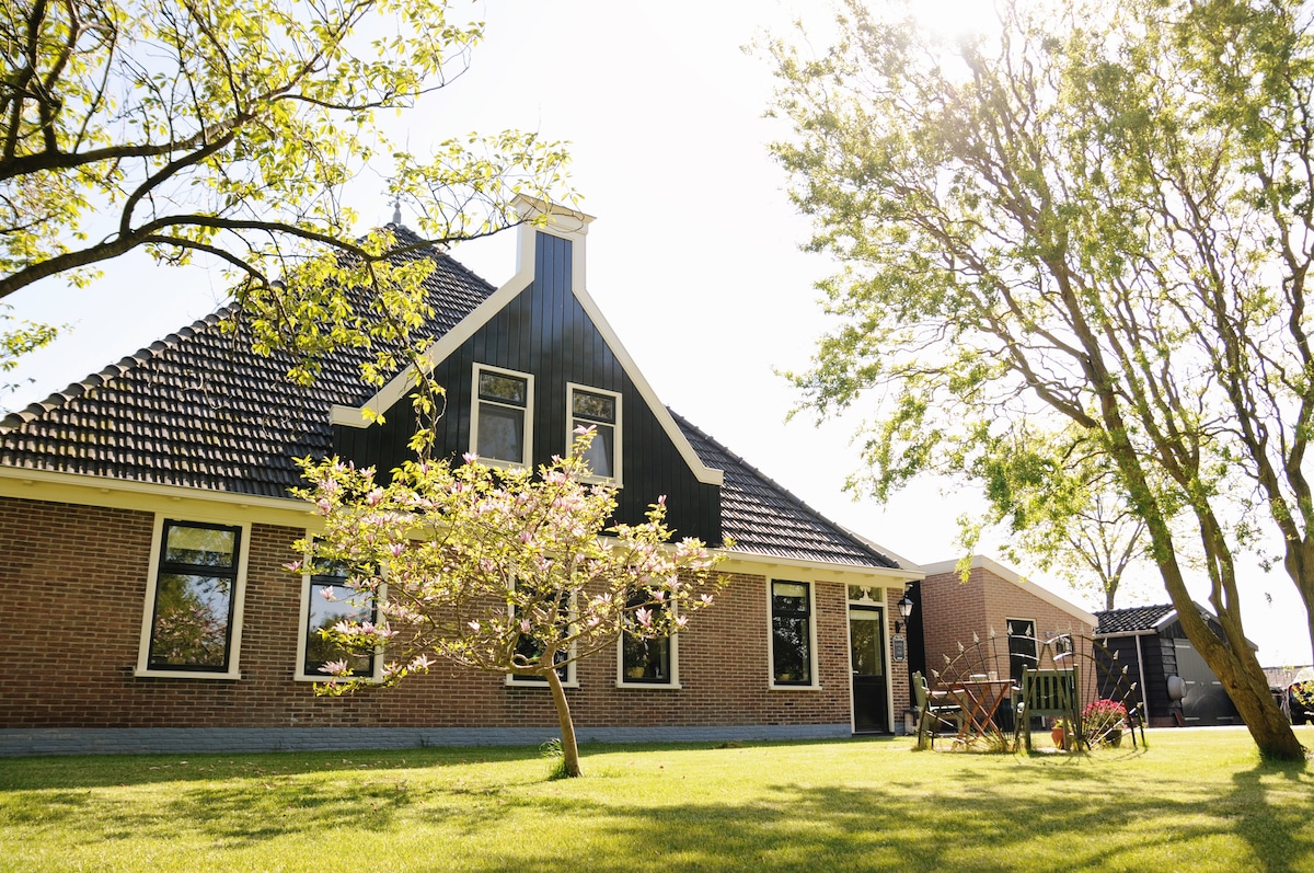A traditional Dutch farmhouse is surrounded by lush greenery, featuring a well-maintained lawn. Sunlight illuminates the structure, highlighting the large windows and asymmetrical roofline. A seating area with chairs is positioned under a tree, offering a serene outdoor space.