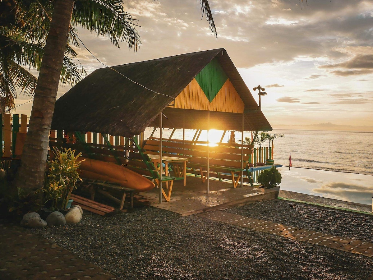 A nipa hut is positioned near the beachfront, showcasing a vibrant green and yellow design. Morning light streams through the open structure, highlighting wooden benches arranged around a table. The gentle waves of the sea can be seen in the background, creating a peaceful atmosphere.