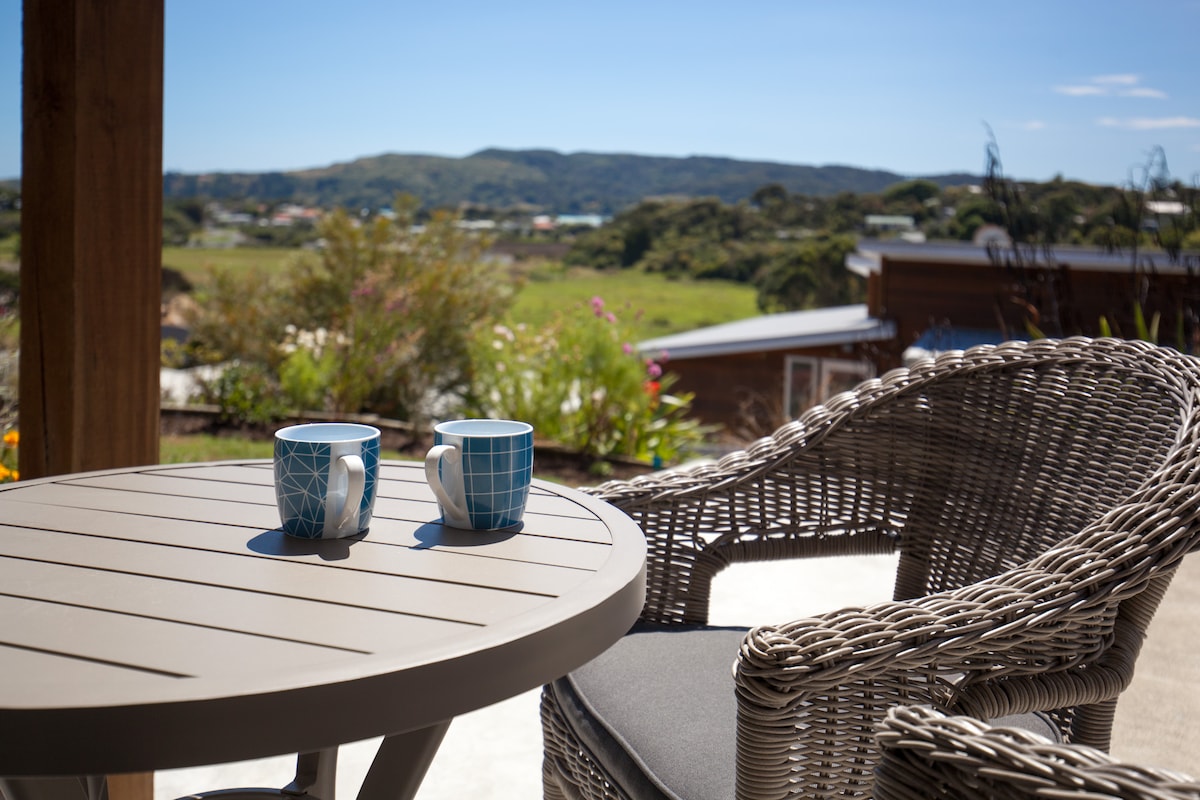 A small outdoor table is set with two patterned mugs, positioned beside a woven chair. The serene landscape extends in the background, featuring rolling hills and greenery under a clear blue sky.