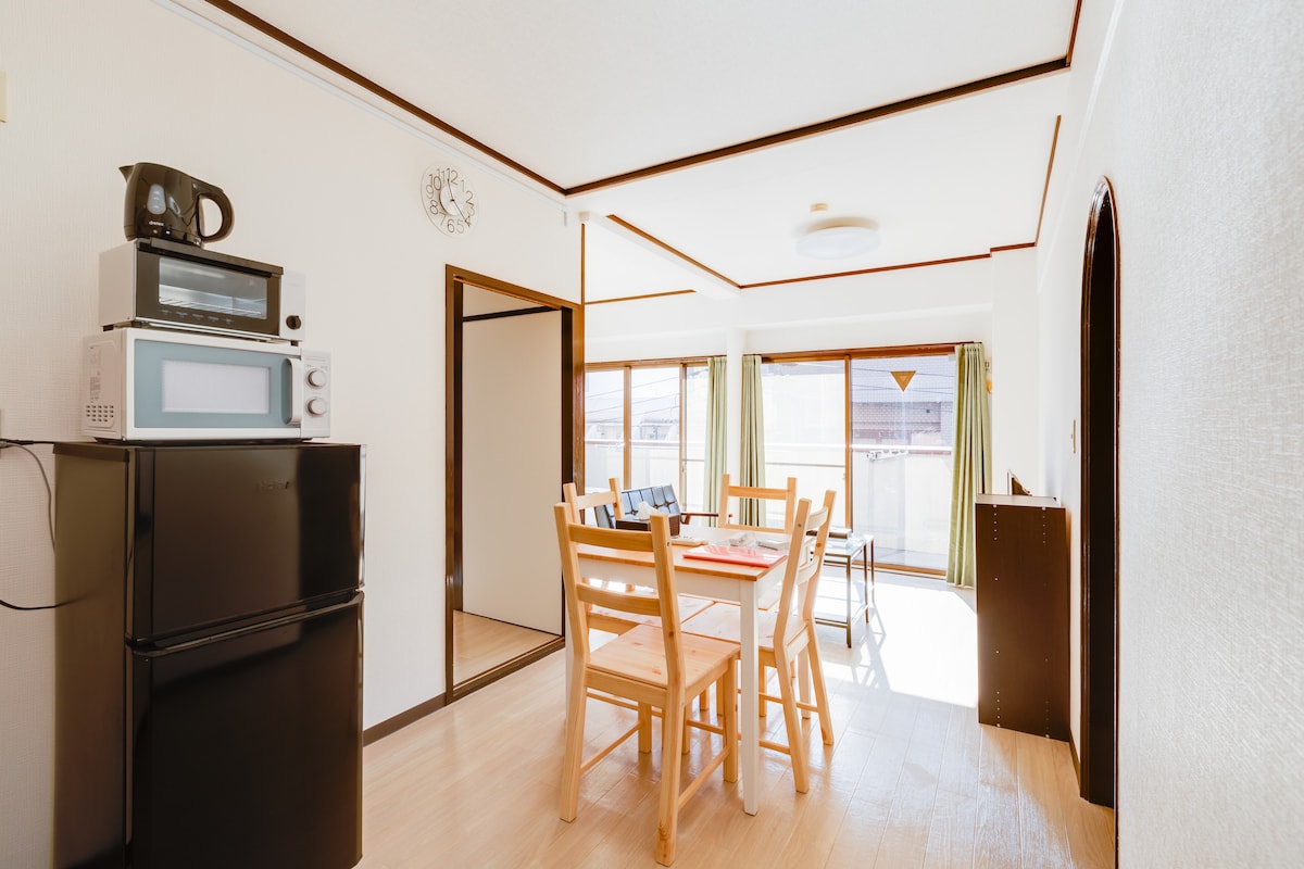 A dining area features a simple wooden table surrounded by chairs, illuminated by natural light. A refrigerator and microwave are visible, along with additional kitchen appliances. Mirrors reflect the bright space, while large windows offer a view of the outdoors.