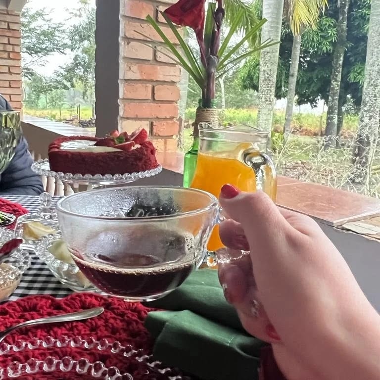 A hand holds a clear cup of coffee, while a red velvet cake and a pitcher of a bright beverage are seen on a table adorned with a vibrant cloth. Lush greenery and palm trees are visible in the background, adding a natural ambiance.