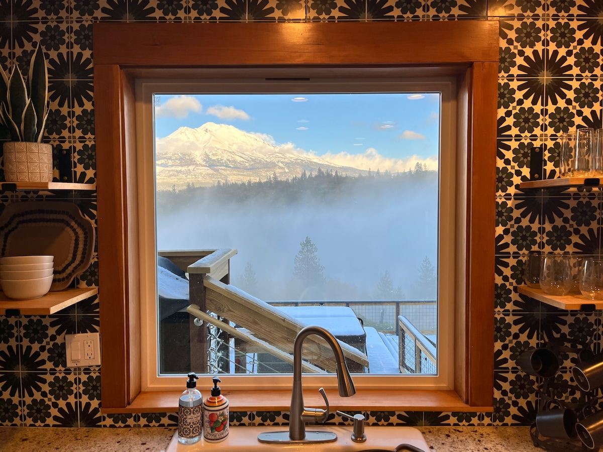 A large window frames a stunning view of snow-covered Mount Shasta, with clouds hovering below. The kitchen sink and modern faucet are positioned in front of the window, while patterned tiles and wooden shelves add a warm touch to the space.