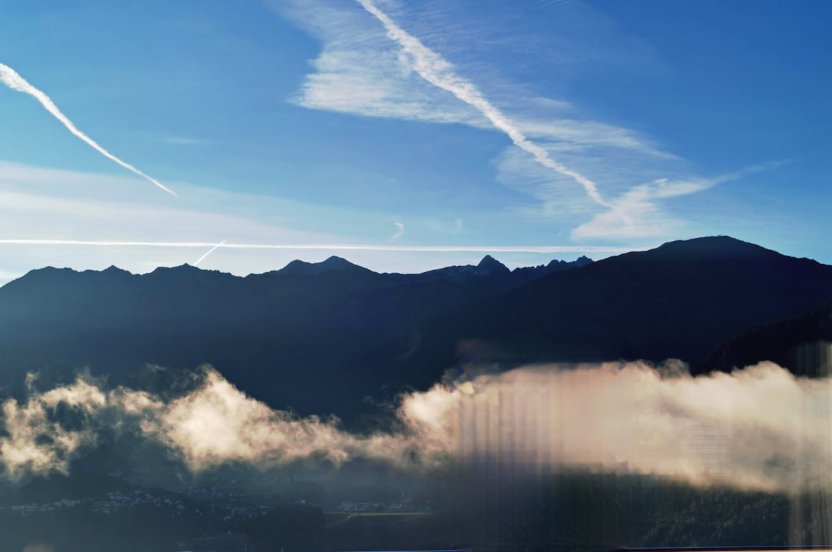 A serene mountain landscape is captured under a clear blue sky, with soft clouds lingering in the valleys. The silhouette of rugged mountains is visible in the distance, creating a tranquil and natural backdrop.