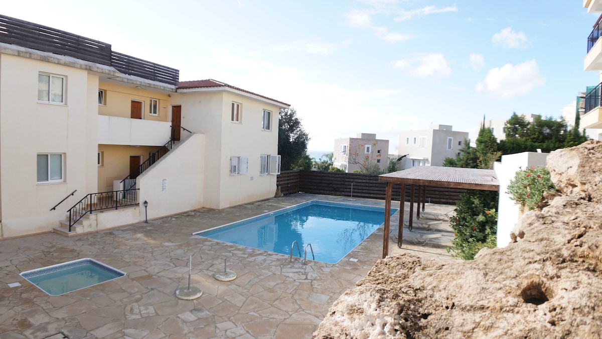 A communal swimming pool is set within a landscaped area of the apartment complex. The pool is surrounded by stone tiles, with a smaller jacuzzi nearby. Two levels of buildings are visible in the background, showcasing simple and modern architectural features.