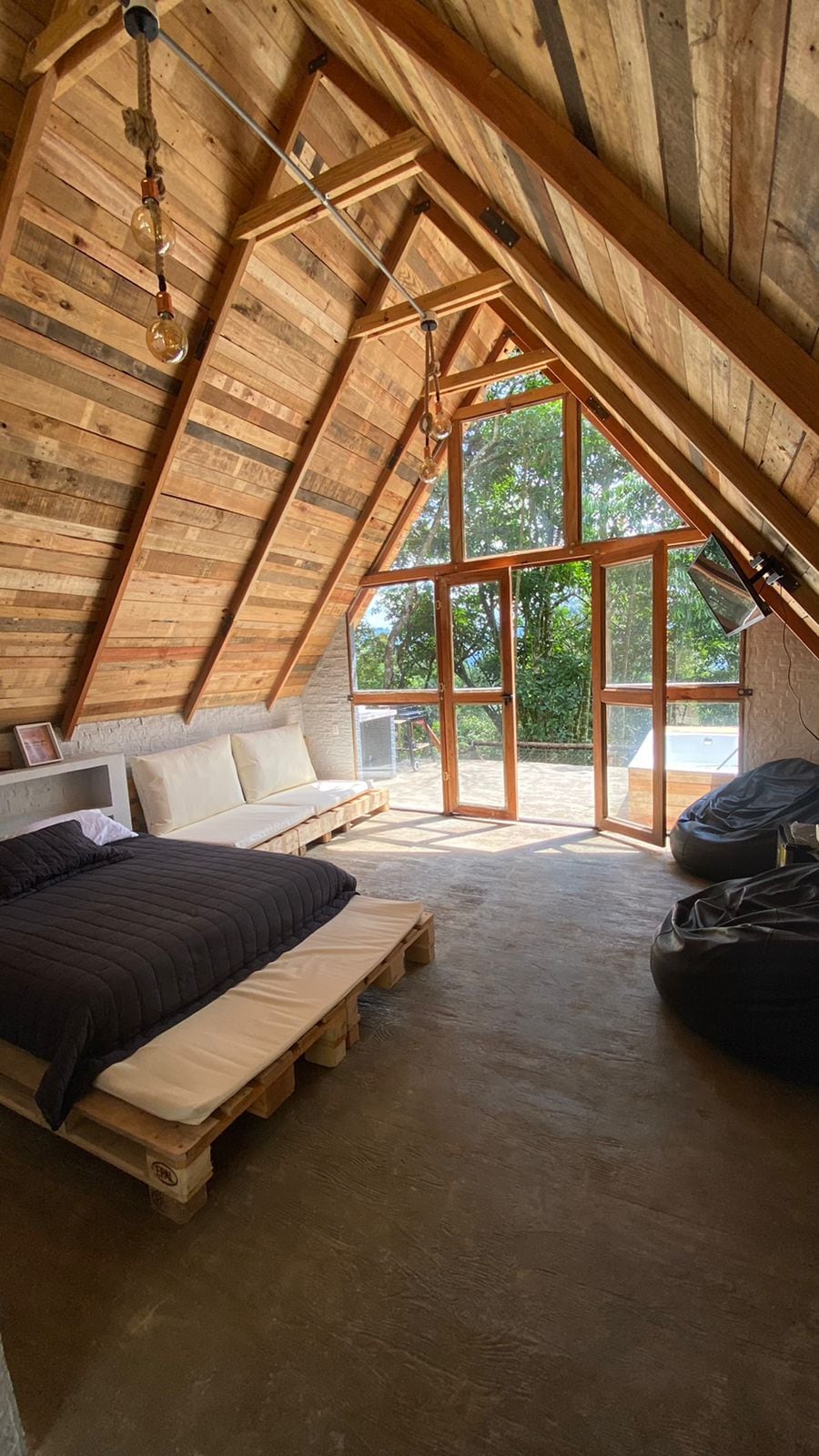 An inviting sleeping area features a spacious bed made of pallets covered in dark linens. Natural light floods through large windows, illuminating the wooden walls and ceiling. Cozy bean bag chairs and additional seating are arranged nearby, creating a relaxed atmosphere.
