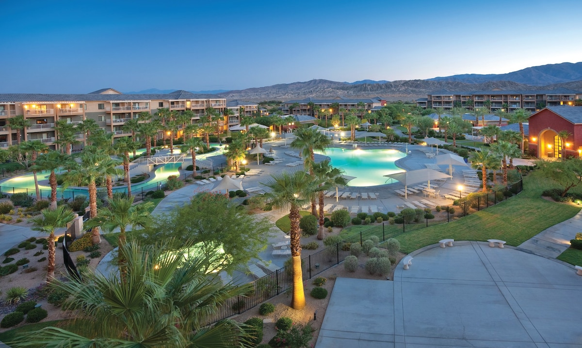 The resort is displayed in a panoramic view, featuring landscaped grounds with palm trees and serene pools illuminated by soft lighting. The surrounding mountains create a natural backdrop, while the modern architecture of the buildings adds a contemporary touch.