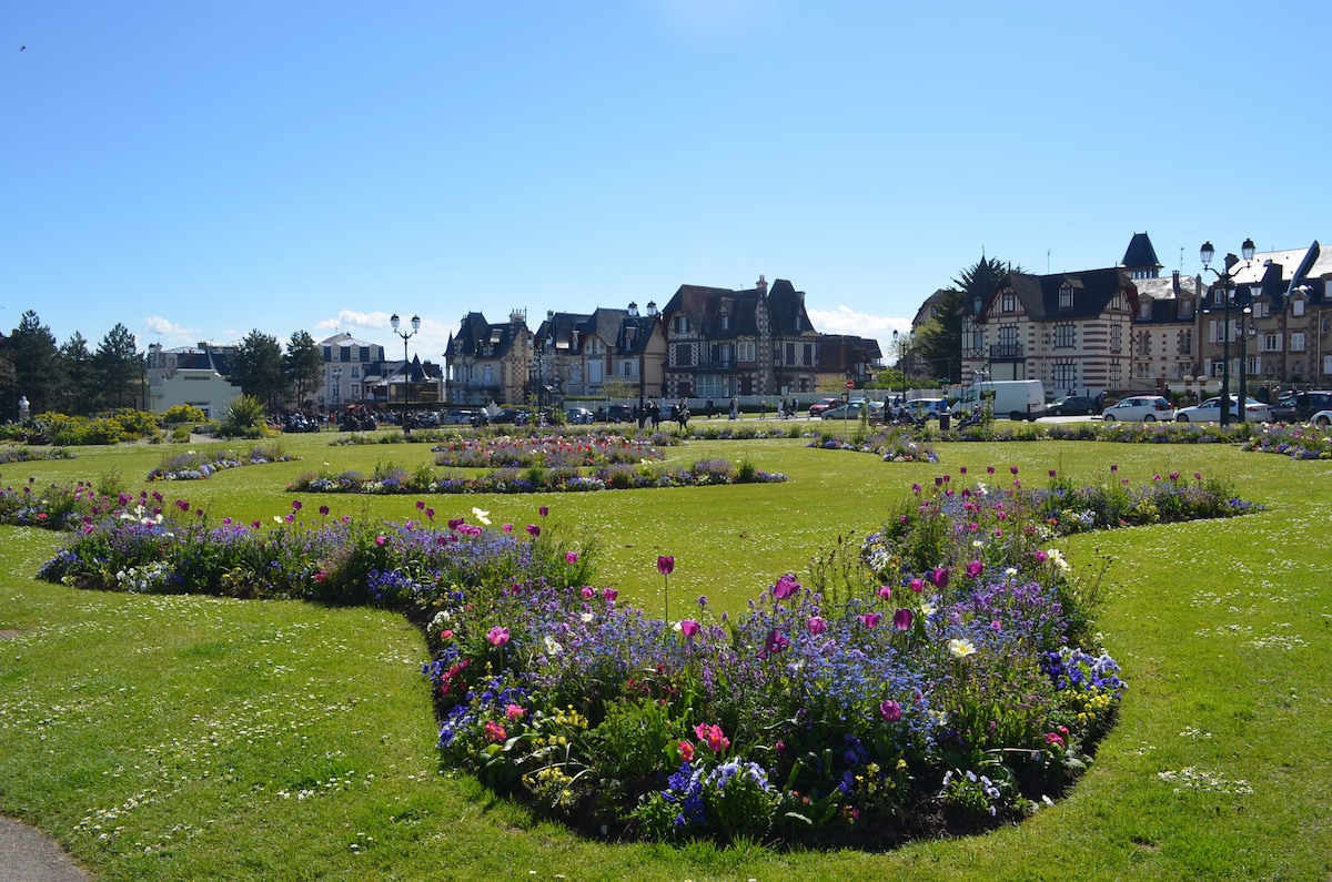 A landscaped garden is set against a clear blue sky, featuring vibrant flower beds in various colors. Neatly trimmed grass surrounds the floral displays, while charming buildings line the background, hinting at nearby amenities and a welcoming atmosphere.