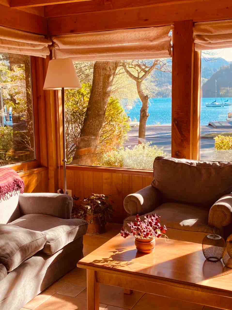 A light-filled sitting area features two gray armchairs beside a wooden coffee table. Large windows showcase views of the lake and lush greenery, while a lamp and potted plants add warmth to the inviting space.