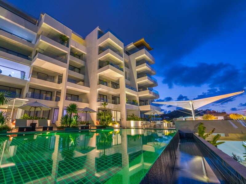 A modern building is showcased against a twilight sky, with multiple balconies and large windows reflecting the water's surface from an infinity pool. Surrounding greenery and shaded lounge areas enhance the ambiance, creating a serene environment for relaxation.