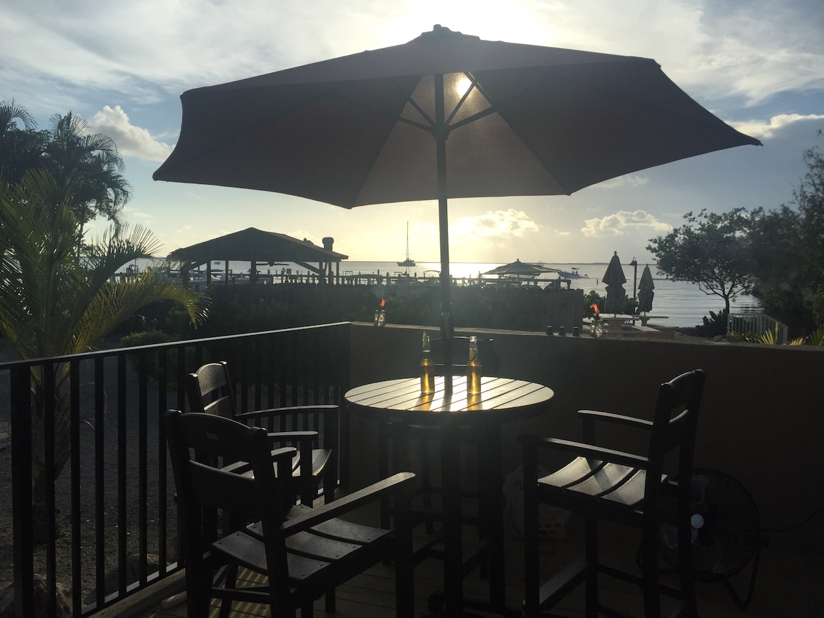 A shaded outdoor patio features a round table surrounded by four tall chairs, overlooking the water. The scene captures the warm glow of the setting sun, with glimpses of palm trees and umbrellas in the background, creating a serene atmosphere.
