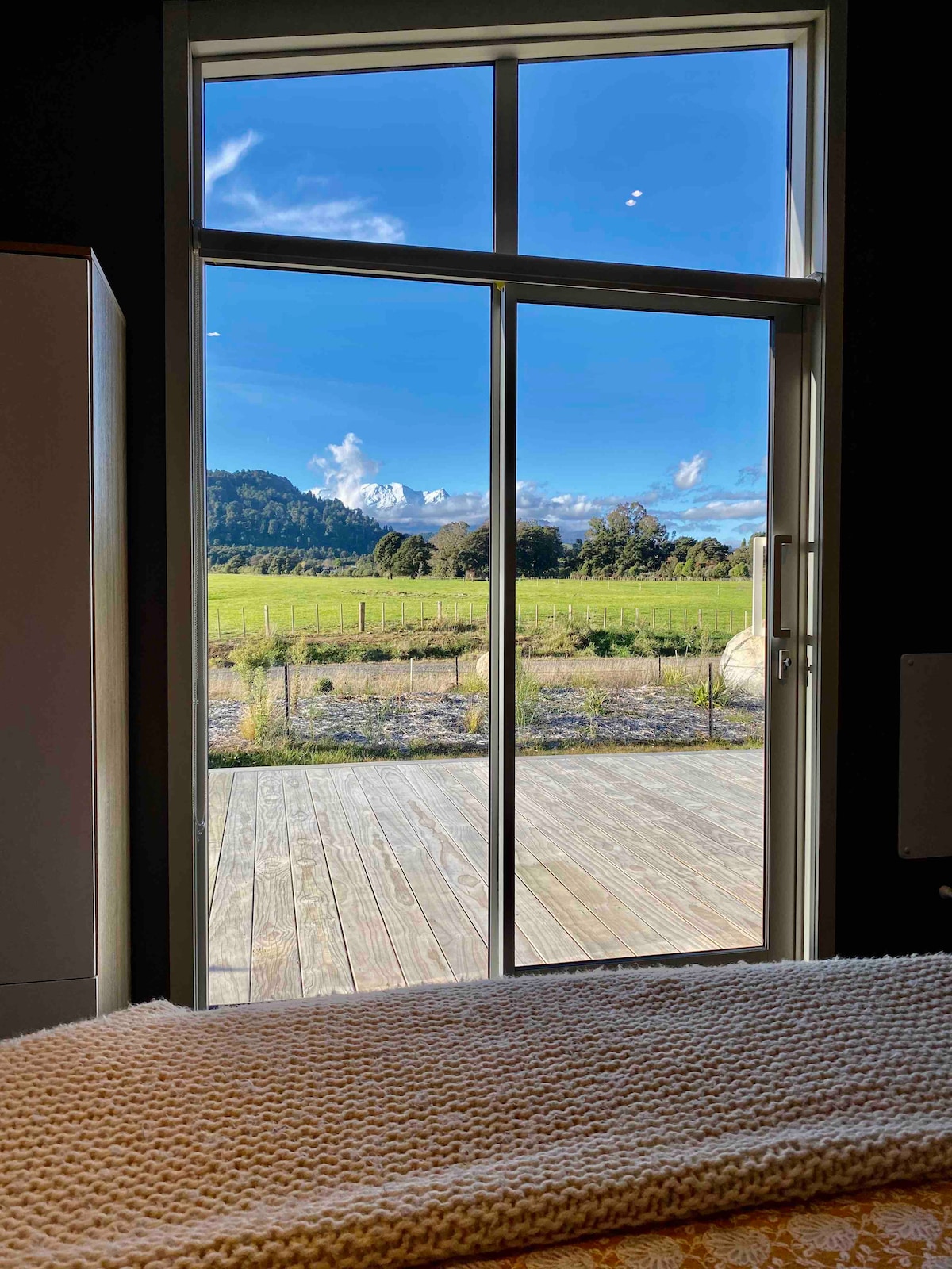 A large glass door opens to expansive countryside views, showcasing green fields and distant hills under a bright blue sky. Natural light fills the space, enhancing the serene atmosphere of the bedroom.