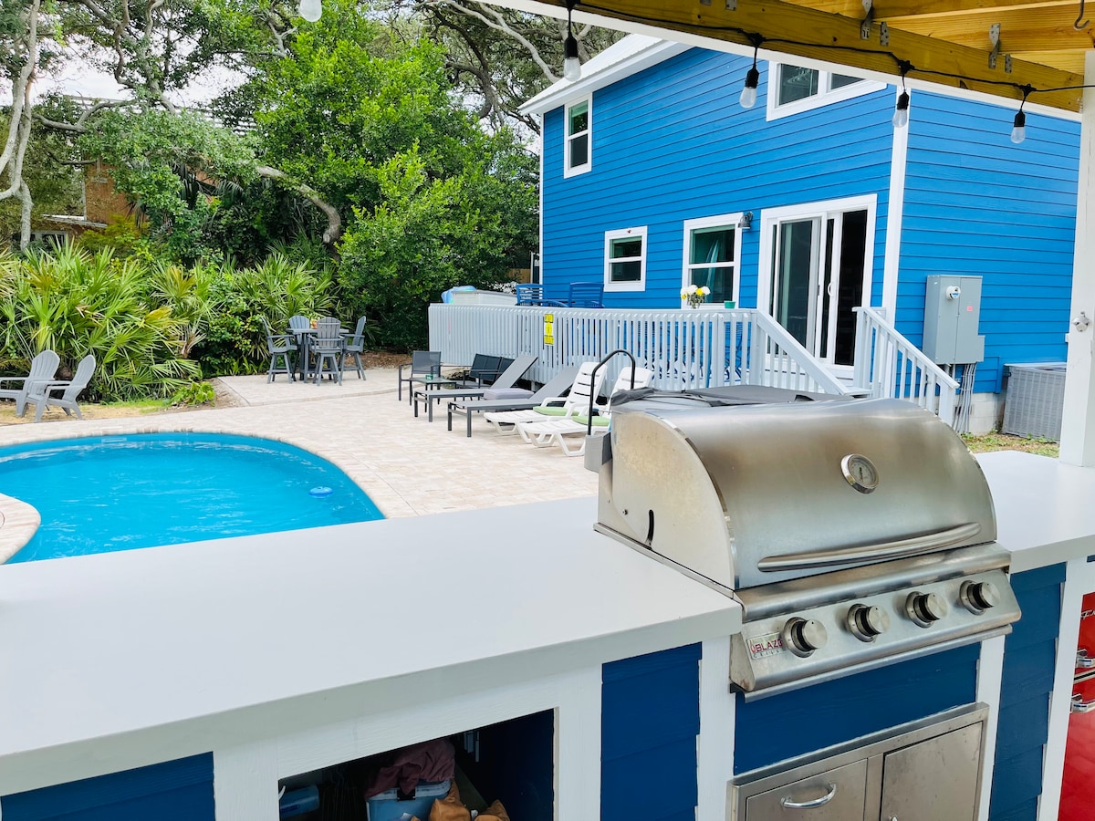 An outdoor space features a sparkling blue pool surrounded by lounge chairs and a patio area. A covered outdoor bar area showcases a gas grill, while lush greenery and palm plants enhance the tropical ambiance. The blue house is visible in the background.