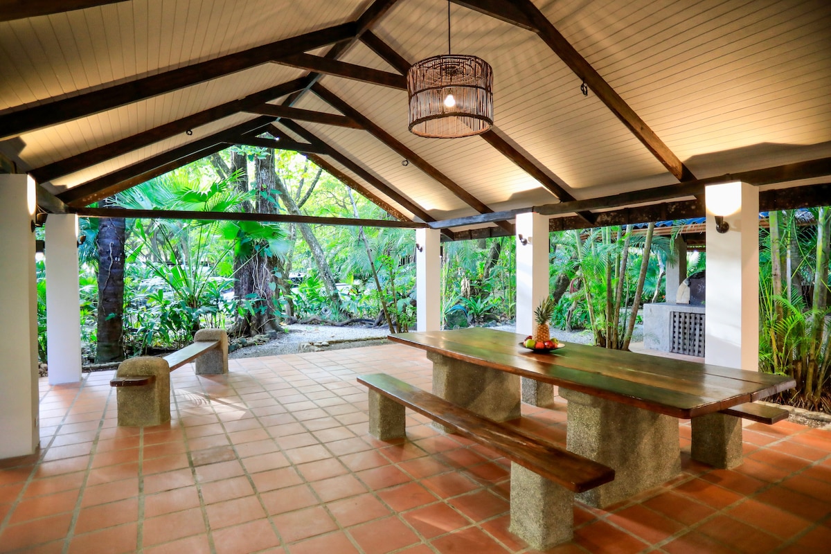 An open-air kitchen and community gathering space is highlighted, featuring a long wooden table flanked by stone benches. The structure boasts high ceilings with wooden beams and a hanging light fixture. Lush greenery surrounds the area, creating a natural backdrop.