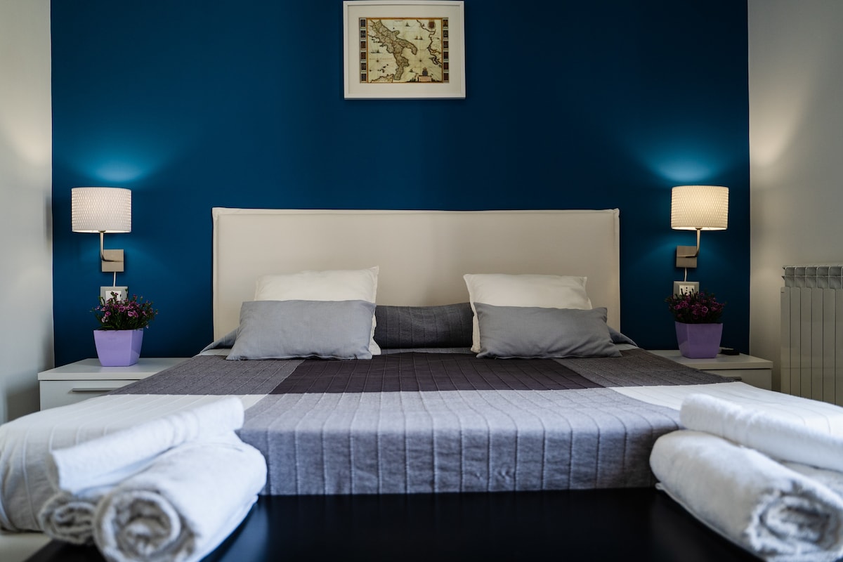 A spacious bed with neutral-toned bedding is centered against a deep blue wall. Soft light is provided by two bedside lamps on nightstands featuring potted flowers. Freshly folded white towels are neatly arranged at the foot of the bed, creating an inviting and tidy appearance.