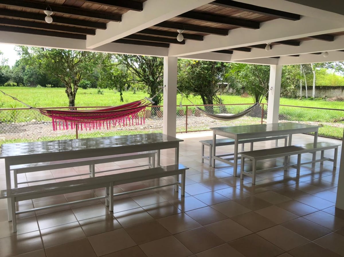 A spacious outdoor area features long tables with benches, providing ample seating for gatherings. Colorful hammocks are visible, suspended between posts, with lush green fields beyond the fence. The ceiling has exposed beams, and bright natural light illuminates the space.