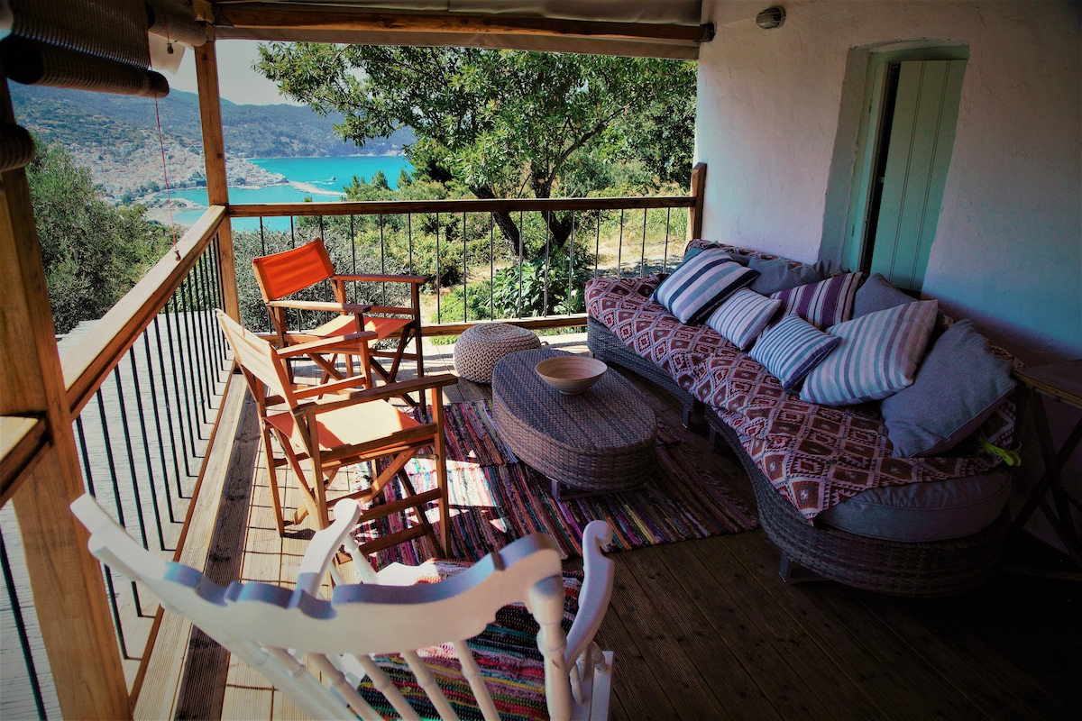 A cozy outdoor seating area features a unique curved sofa adorned with patterned cushions, alongside wooden chairs in orange and white. A round table sits in the center, all positioned on a rustic wooden deck with a vibrant area rug, offering views of the surrounding landscape and water.