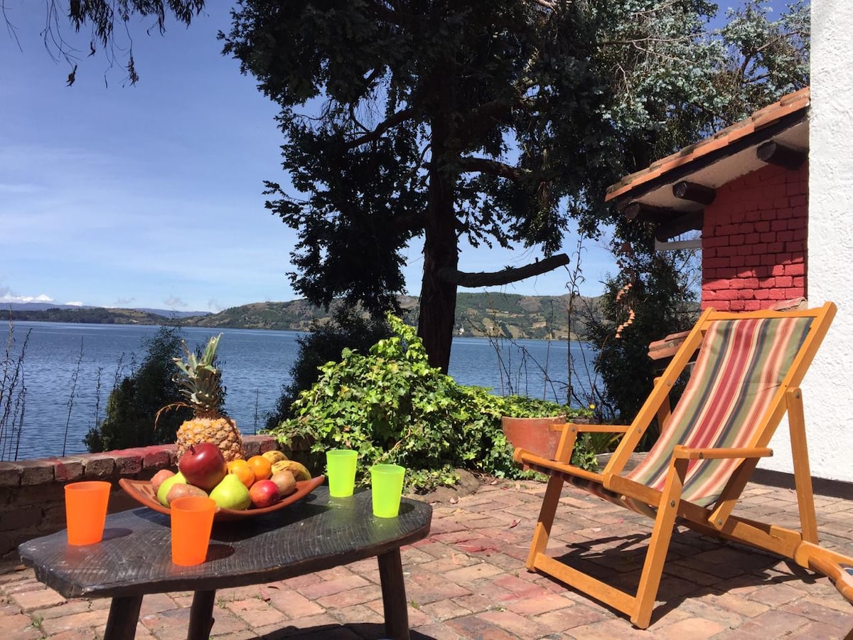 An outdoor terrace presents a relaxed seating area with a wooden chair and a table. A bowl of fresh fruits, including pineapple and apples, is displayed, while bright cups add a touch of color. The serene view of the lake and surrounding nature enhances the setting.