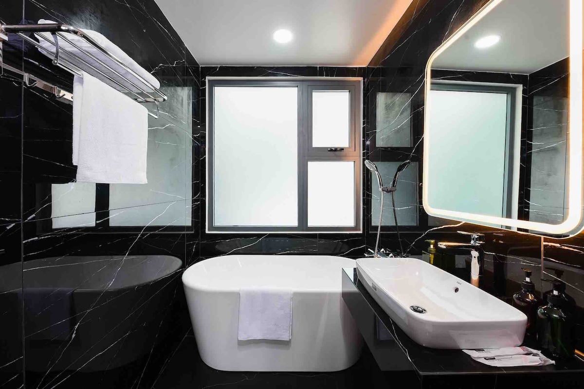 A modern bathroom features a spacious free-standing bathtub and a sleek vanity with a basin. Large windows allow natural light, and elegant marble tiles create a sophisticated ambiance. Soft, white towels are neatly arranged on a rack, complemented by backlit mirror illumination.