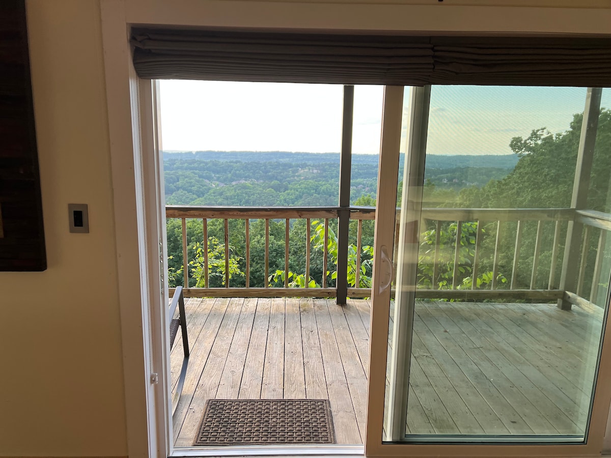 A spacious balcony is visible through glass sliding doors, featuring a wooden deck surrounded by lush greenery. Scenic views of rolling hills and valleys extend into the distance. A welcome mat lies at the entrance, inviting guests to enjoy the outdoor space.