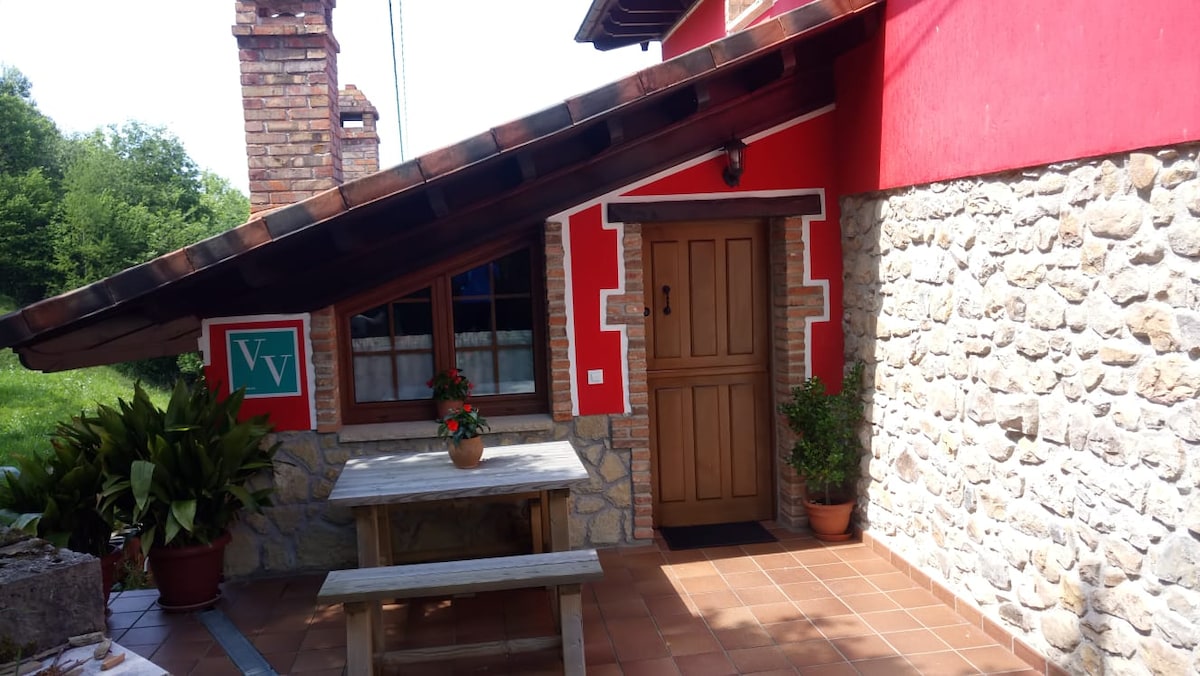 A rustic exterior is showcased with a combination of wood and stone materials. A warmly colored front door is framed by a wood roof structure. A wooden table and potted plants are placed on the tiled patio, inviting relaxation in the surrounding natural setting.
