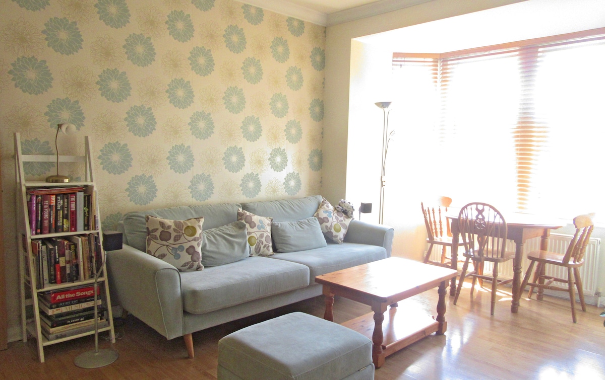 A light-filled living area is characterized by a large, comfortable sofa and a wooden coffee table. A bookshelf displays an assortment of books, while a dining table for four is positioned by the window, framed by decorative floral wallpaper.