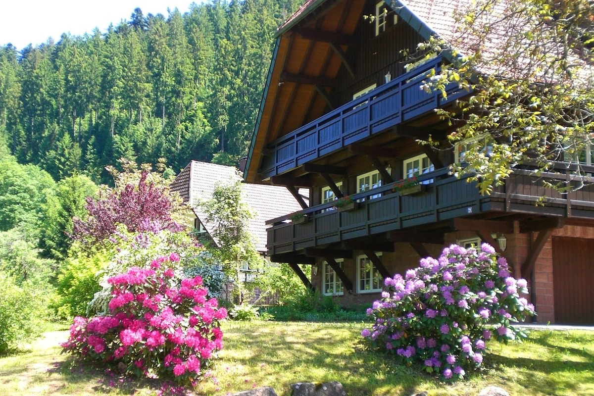 A garden featuring vibrant pink and purple flowers is situated in front of a multi-level wooden structure. The building's balconies overlook the greenery, surrounded by trees that provide a natural backdrop. The scene reflects a serene and peaceful countryside atmosphere.