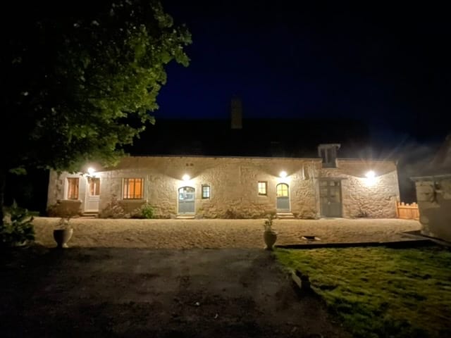 The exterior of the countryside house is illuminated at night, showcasing its rustic stone façade. Soft lighting accentuates the entrance, highlighting the various doorways and windows. Surrounding trees provide a natural backdrop, while a gravel pathway leads to the welcoming facade.