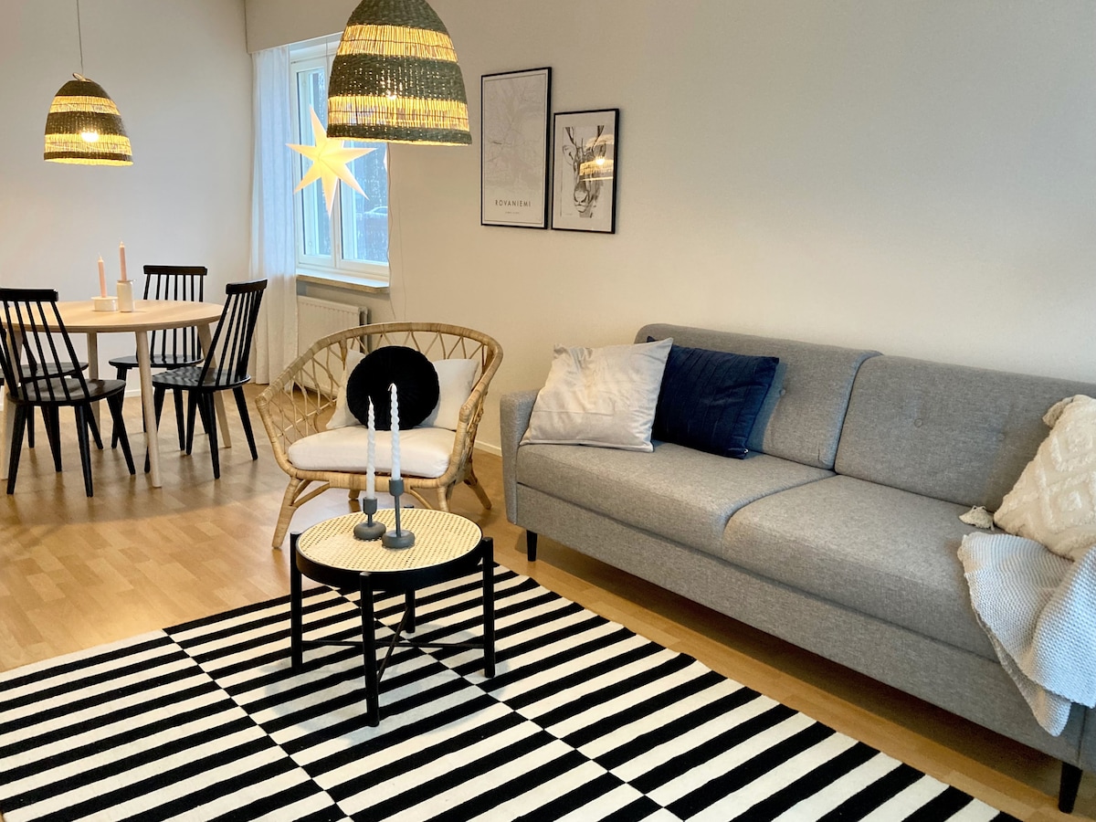 A bright living area features a gray sofa paired with a patterned black and white rug. A coffee table sits in front, adorned with candles. In the background, a dining table for four is surrounded by black chairs, complemented by pendant lighting overhead.