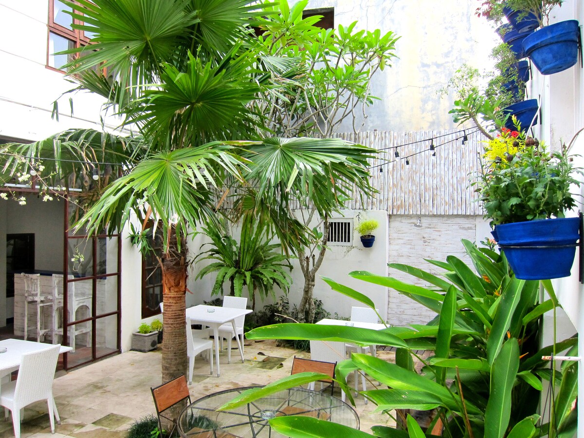 A serene courtyard is filled with lush tropical plants and trees. Brightly colored flower pots hang from the walls. Tables and chairs are set among the greenery, offering a calm space for relaxation. String lights are draped between the trees, adding a welcoming touch.