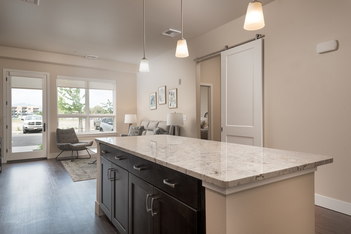 An open living space showcases a modern kitchen island with dark cabinetry and a speckled countertop. Pendant lights hang above, illuminating the area. In the background, a cozy seating area features framed artwork, a light gray sofa, and natural light from large windows.