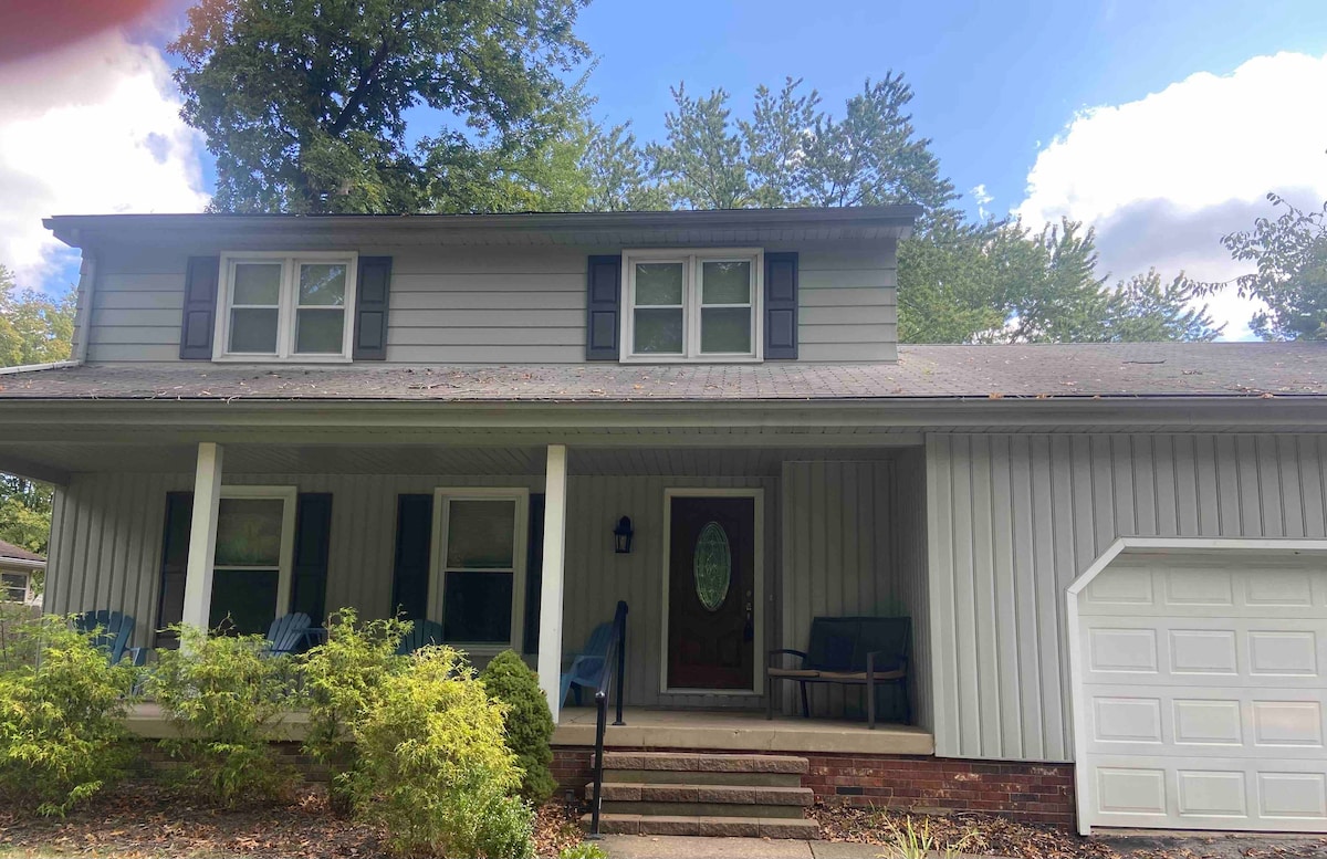 The home features a welcoming front porch with two blue chairs and a small table, surrounded by greenery. The two-story structure has a gray exterior with dark shutters and a prominent entry door. A single-car garage is located to the right.