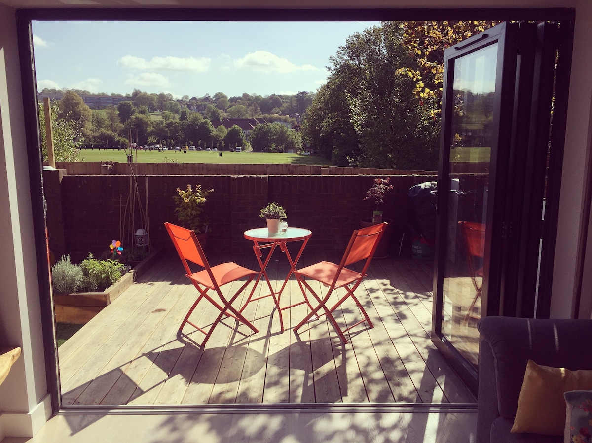A wooden deck area features a small round table accompanied by two bright chairs. Greenery is visible in planters, with a view of open fields and trees in the distance through bi-folding doors that provide ample natural light.