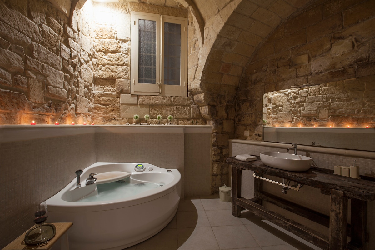 A spacious bathroom is highlighted by a two-person hydromassage tub, set against rustic stone walls. An antique wooden workbench serves as a sink area, complemented by soft lighting from tea candles arranged along the tub's ledge. A window above allows natural light to filter in.