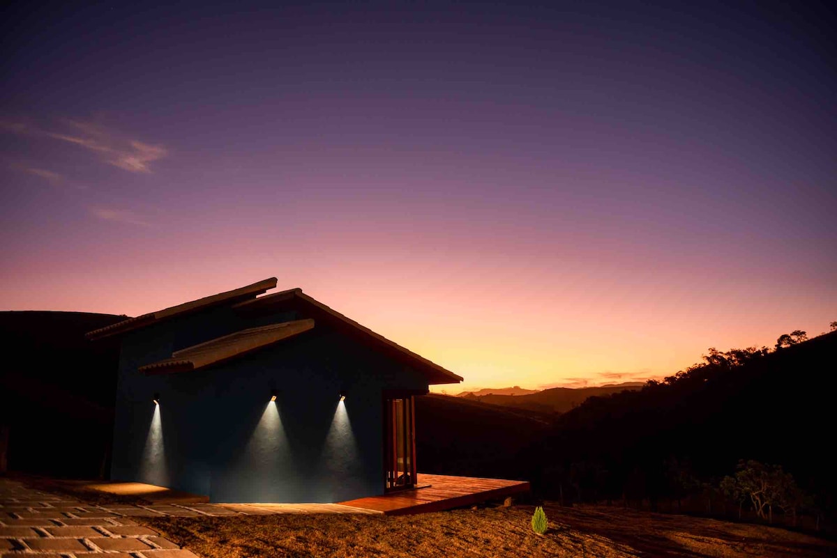 The exterior of the chalé is shown at dusk, featuring a modern design highlighted by exterior lighting. The backdrop reveals a gradient sky transitioning from vibrant orange to deep blue, with mountain silhouettes visible in the distance.