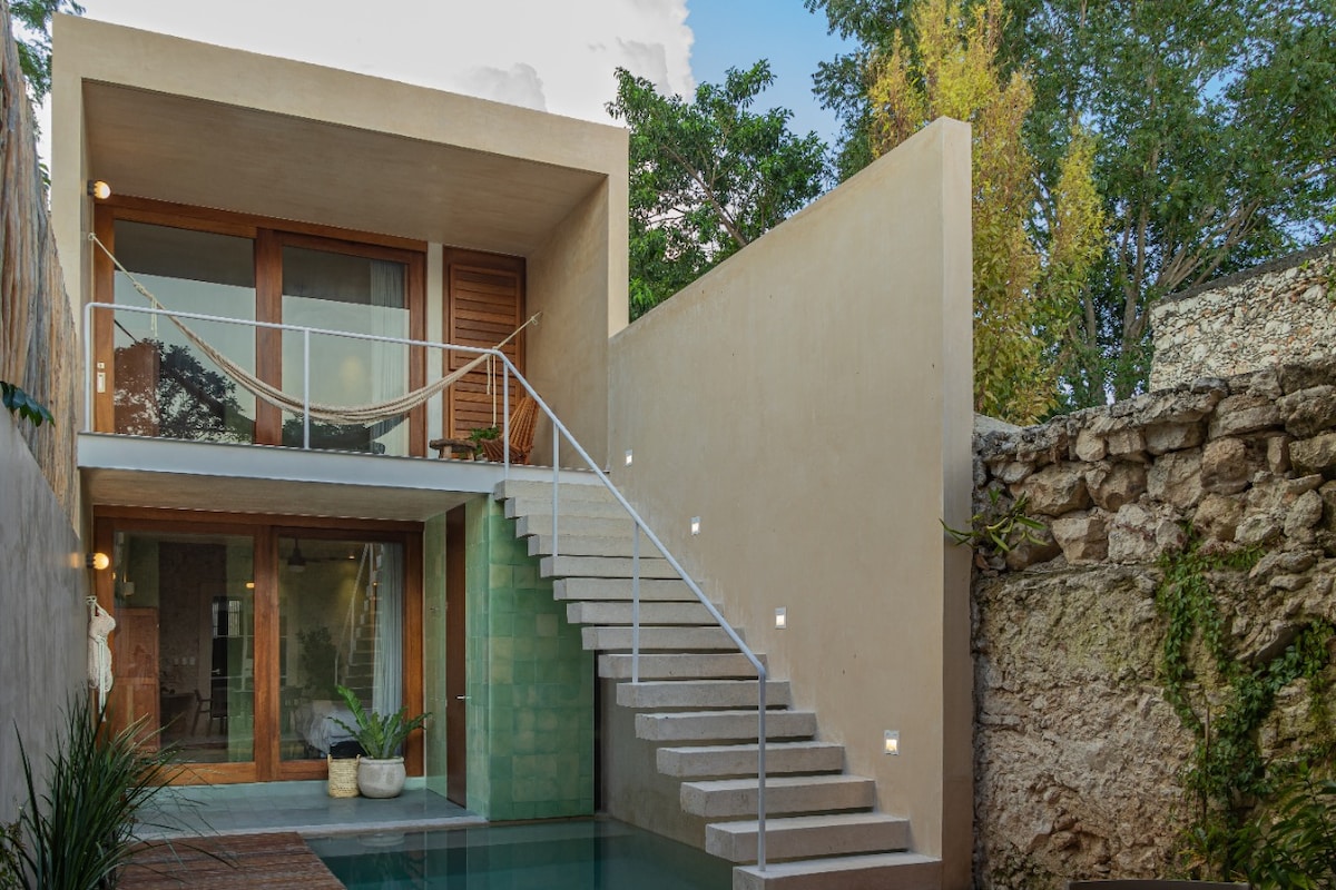 The exterior of the restored house features a modern design with a spacious staircase leading to the upper level. A swimming pool is visible in the foreground, accompanied by plants in decorative pots, while a hammock is strung between two posts for relaxation.