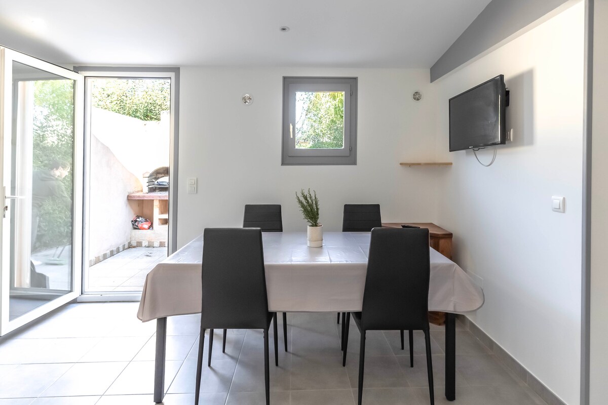 A modern dining area features a rectangular table with six black chairs. Natural light fills the space through large glass doors that open to a patio. A small plant sits at the center of the table, with a wall-mounted television and a shelf positioned nearby.