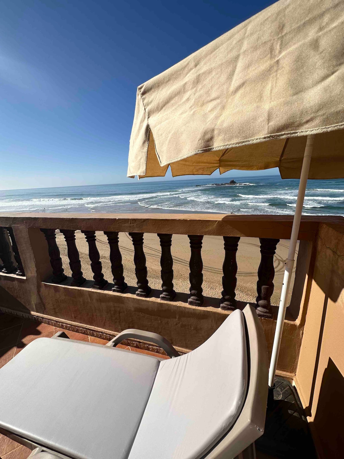 A sun lounger is positioned on a private terrace overlooking the Atlantic Ocean. A sunshade provides shelter from daylight, while gentle waves can be seen lapping against the shore in the background. The railing accentuates the view of the expansive sandy beach.